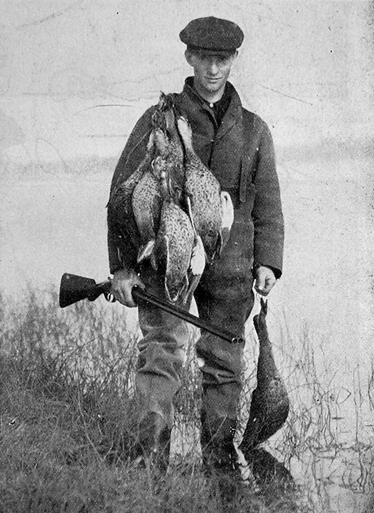 A historic black and white photo of a duck shooter holding a rifle and ducks