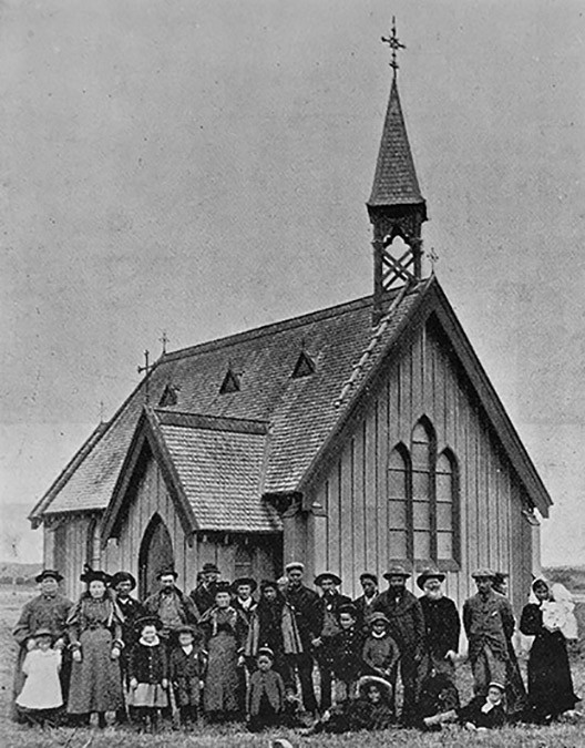 An historic black and white photo of people standing in front of a small church