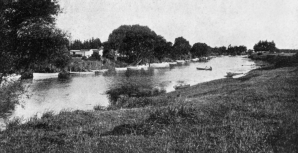 A historic black and white photo of small boats along the mouth of the Waikirikiri/Selwyn River, with some of the Selwyn Huts in the background