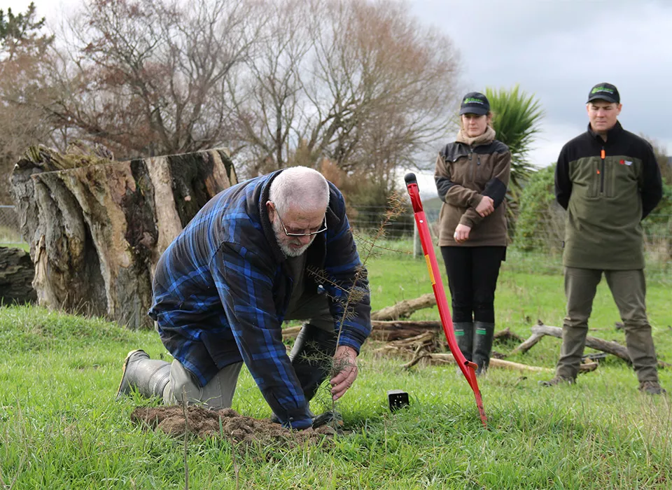 A man in a blue jacket kneeling down and planting a tree at Te Waihora